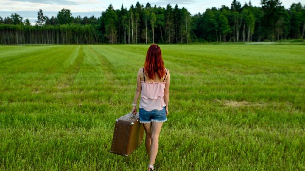 Woman with red hair and vintage suitcase walking through Kolga's lush meadow in Estonia on a summer day.