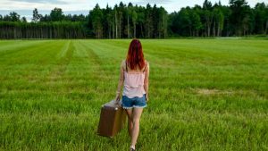 Woman with red hair and vintage suitcase walking through Kolga's lush meadow in Estonia on a summer day.